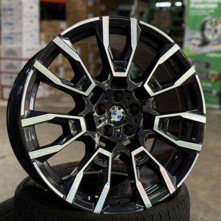 Black and silver BMW alloy wheel close-up, showing intricate spoke design and center badge on a tire in a showroom setting.] ,