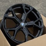 Two black BMW alloy wheels stacked in a cardboard box on a rough asphalt surface.