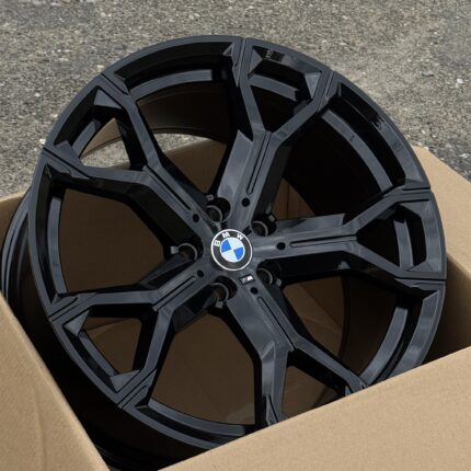 Two black BMW alloy wheels stacked in a cardboard box on a rough asphalt surface.