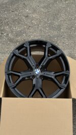 Black BMW alloy wheel with a geometric spoke design in a cardboard shipping box on a gravel surface, viewed from above.