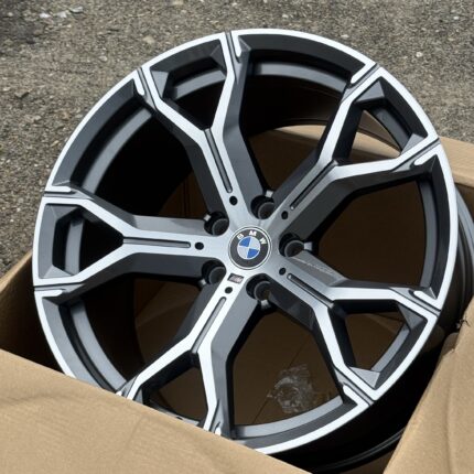Close-up of a black and silver alloy BMW wheel resting in a cardboard box on a gravel surface outside.