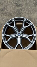 BMW alloy wheel with geometric spokes resting in a cardboard box, front view show logo at center cap (M badge present).