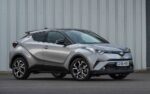 Silver Toyota C-HR compact SUV parked on a paved surface in front of a corrugated metal wall, showing its design and alloy wheels