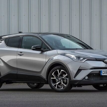 Silver Toyota C-HR compact SUV parked on a paved surface in front of a corrugated metal wall, showing its design and alloy wheels