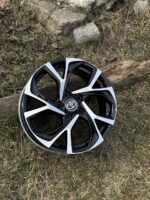 Black and white alloy wheel rim with a Toyota emblem resting on dry grass outdoors on rocks and wood nearby.