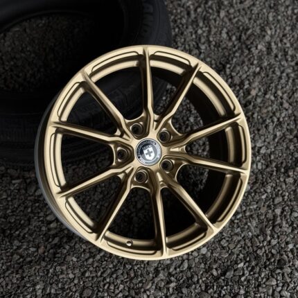 Gold-colored alloy wheel with intricate multi-spoke design resting on gravel, center cap visible.