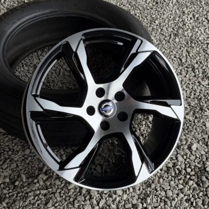 Five-spoke silver and black Volvo alloy wheel with center cap, lay on a gravel surface beside a tire