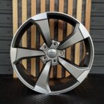 Audi alloy wheel rim with a five-spoke design and central logo, standing upright against a wooden backdrop.
