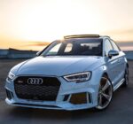White Audi RS5 parked on a rooftop at sunset, front grille and alloy wheels visible.