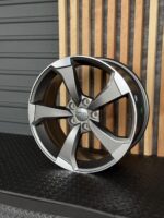 Five-spoke silver Audi alloy wheel leaning against wooden slats on a black textured floor, hub with Audi logo visible