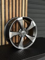 Audi alloy wheel with five split spokes resting on a textured floor beside a wooden slat wall, angled to show the front and logo hub.