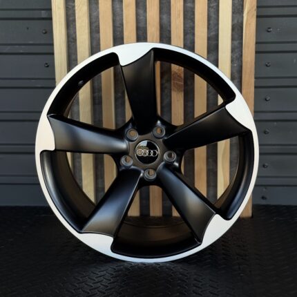 Black five-spoke alloy wheel with Audi center cap and white outer lip, propped against a wooden and metal backdrop.