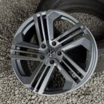 Volkswagen alloy wheel with a multi-spoke design resting on small gray gravel, tire visible in the background on the right-side face of the wheel.