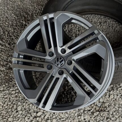 Volkswagen alloy wheel with a multi-spoke design resting on small gray gravel, tire visible in the background on the right-side face of the wheel.