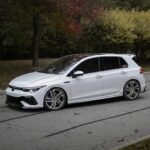 White Volkswagen hatchback with lowered suspension and large multi‑spoke wheels parked on a street with autumn foliage in the background, side windows tinted.