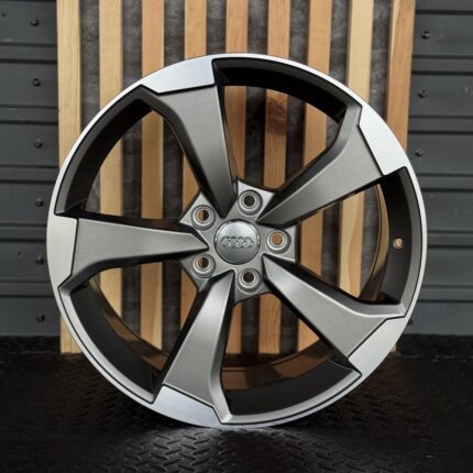 Audi alloy wheel rim with five split spokes and central Audi logo, set against a wooden backdrop.