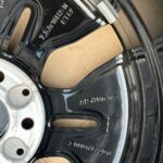 Close-up of a glossy black alloy car wheel rim showing embossed part numbers and openings for lug bolts that reveal the hub area.
