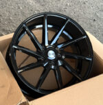 Black glossy multi-spoke alloy car wheel resting in a cardboard box on a rough pavement surface.