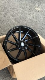 Glossy black multi-spoke car alloy wheel in a cardboard box on a gravel surface