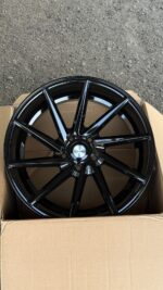 Black, glossy multi-spoke alloy wheel (10 spokes) with a chrome center cap reading 'Vossen' inside an open cardboard box on a gravel surface.