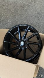 Glossy black multi-spoke car wheel housed in a cardboard box on an asphalt surface, ready for installation.