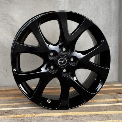 Black glossy alloy wheel rim with Mazda emblem at the center cap, resting on a wooden surface.