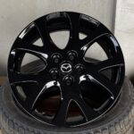 Close-up of a glossy black Mazda alloy wheel with 10 curved spokes and center cap logo, mounted on a tire against a concrete backdrop