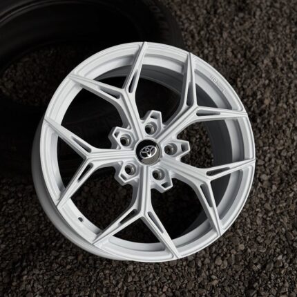 White alloy wheel with a Toyota center cap resting on gravel, showcasing a modern multi-spoke design.