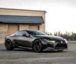 Black Lexus LC 500 coupe parked on a gravel lot near a beige industrial building at dusk.