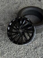 Black glossy alloy car wheel with Toyota center cap lying on gravel beside a tire on the ground, ready for mounting.