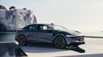 Side view of a sleek gray sedan parked on a white display platform by the sea, with white cliffs in the background.