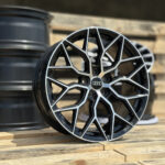 Close-up of a black and silver multi-spoke Audi alloy wheel resting on a wooden pallet surface on a sunny day.