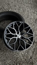 Close-up of a glossy alloy wheel with a geometric silver-and-black spokes design, centered by a Vossen cap, resting near a tire on gravel.