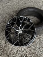 Black glossy Vossen alloy wheel with a complex geometric spoke pattern resting on a gravel surface beside a tire.