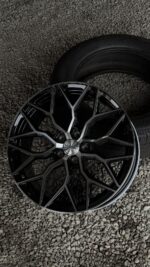 Close-up of a glossy black Vossen alloy wheel with a geometric multi-spoke design on gravel, with a tire in the background.