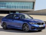 Blue BMW sedan parked on pavement in front of a modern glass-front building, angled view showcasing front grille and alloy wheels