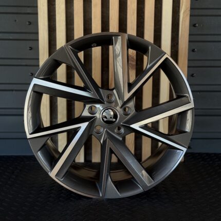 Front view of a dark gray multi-spoke alloy wheel with silver accents and a Skoda center cap on a textured surface.