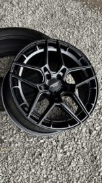 Black multi-spoke alloy wheel on a gravel surface, center cap showing a brand logo.