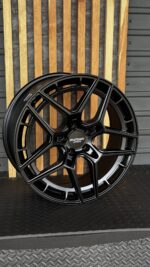 Black multi-spoke forged alloy wheel with center cap reading 'Forged wheels' against a wooden backdrop.