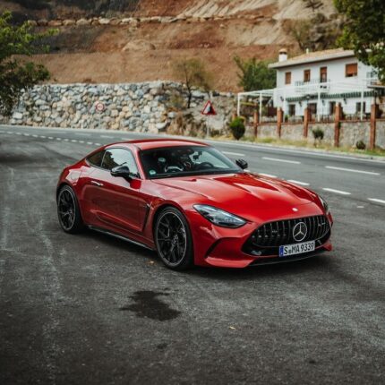 Red Mercedes AMG GT-style sport coupe parked on a winding road beside a stone wall and houses in the background.