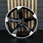 Black five-spoke Audi alloy wheel with a white outer rim displayed upright, center cap with Audi logo visible.