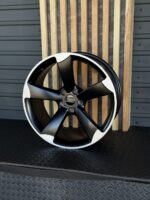 Black and white five-spoke Audi alloy wheel leaning against vertical wooden slats on a dark floor.