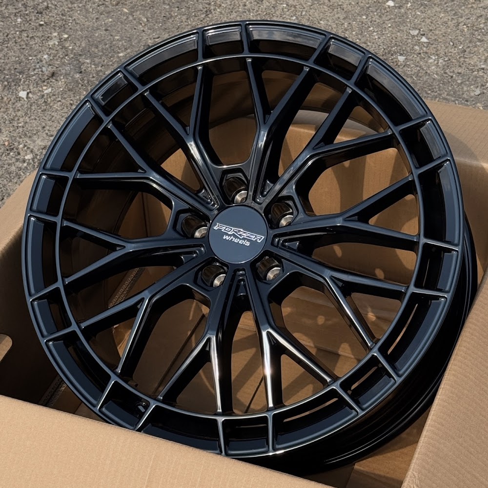 Screenshot Close-up of a glossy black alloy wheel with an intricate multi-spoke design, resting in a cardboard box, center cap reads 'Forged wheels'.