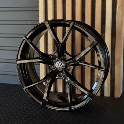 Black glossy alloy wheel with a Volkswagen emblem center, standing against wooden slats.