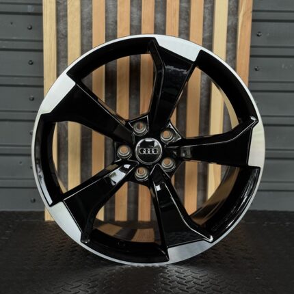 Black glossy five-spoke alloy wheel with Audi center cap displayed against a wooden slat background, close-up view