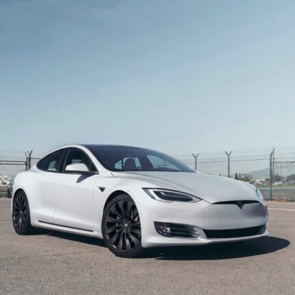White Tesla Model S sedan parked on pavement with a chain-link fence and clear blue sky in the background