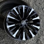 Black multi-spoke alloy wheel with white accents and a Toyota emblem at the center, mounted on a tire on gravel surface.