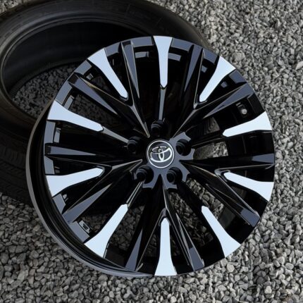 Black multi-spoke alloy wheel with white accents and a Toyota emblem at the center, mounted on a tire on gravel surface.