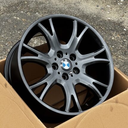 BMW alloy wheel with blue-and-white logo in a cardboard box on a gravel surface.