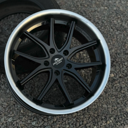 Black multi-spoke forged alloy wheel with a silver lip on a gravel surface, center cap reads Forged Wheels.